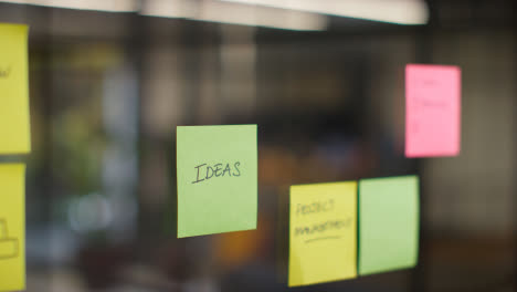 Close-Up-Of-Woman-Putting-Sticky-Note-With-Ideas-Written-On-It-Onto-Transparent-Screen-In-Office-1