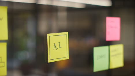 Close-Up-Of-Woman-Putting-Sticky-Note-With-AI-Written-On-It-Onto-Transparent-Screen-In-Office-1