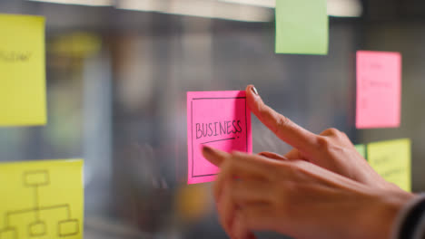 Close-Up-Of-Woman-Putting-Sticky-Note-With-Business-Written-On-It-Onto-Transparent-Screen-In-Office-2