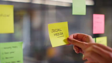 Close-Up-Of-Woman-Putting-Sticky-Note-With-Social-Media-Written-On-It-Onto-Transparent-Screen-In-Office-2