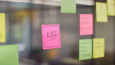 Close-Up-Of-Woman-Putting-Sticky-Note-With-ROI-Written-On-It-Onto-Transparent-Screen-In-Office-1