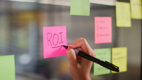 Close-Up-Of-Woman-Writing-ROI-Onto-Sticky-Note-Stuck-To-Transparent-Screen-In-Office-1