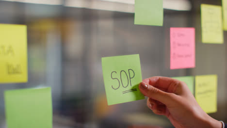 Close-Up-Of-Woman-Putting-Sticky-Note-With-SOP-Written-On-It-Onto-Transparent-Screen-In-Office-2