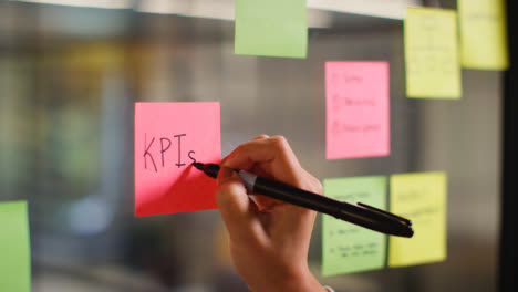 Close-Up-Of-Woman-Writing-KPIs-Onto-Sticky-Note-Stuck-To-Transparent-Screen-In-Office-1