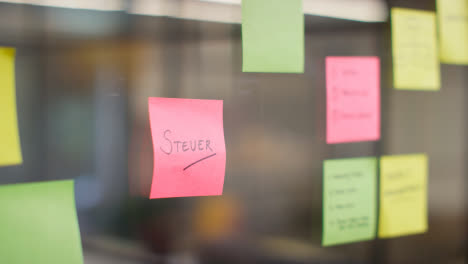 Close-Up-Of-Woman-Putting-Sticky-Note-With-German-Word-Steuer-Or-Tax-Written-On-It-Onto-Transparent-Screen-In-Office-1