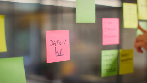 Close-Up-Of-Woman-Putting-Sticky-Note-With-German-Word-Daten-Or-Data-Written-On-It-Onto-Transparent-Screen-In-Office-1