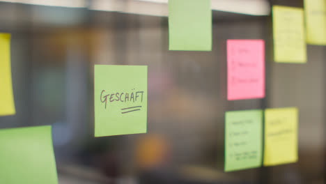 Close-Up-Of-Woman-Putting-Sticky-Note-With-German-Word-Geschaft-Or-Shop-Written-On-It-Onto-Transparent-Screen-In-Office-1