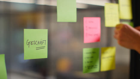 Close-Up-Of-Woman-Putting-Sticky-Note-With-German-Word-Geschaft-Or-Shop-Written-On-It-Onto-Transparent-Screen-In-Office-2