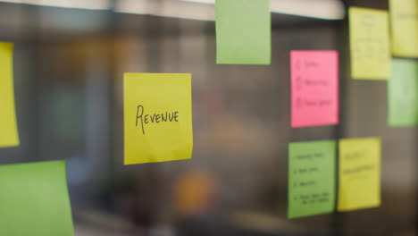 Close-Up-Of-Woman-Putting-Sticky-Note-With-Revenue-Written-On-It-Onto-Transparent-Screen-In-Office-1