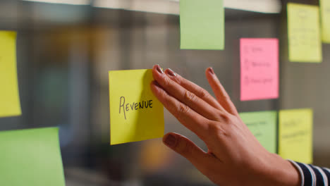 Close-Up-Of-Woman-Putting-Sticky-Note-With-Revenue-Written-On-It-Onto-Transparent-Screen-In-Office-2