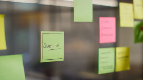 Close-Up-Of-Woman-Putting-Sticky-Note-With-Start-Up-Written-On-It-Onto-Transparent-Screen-In-Office-1