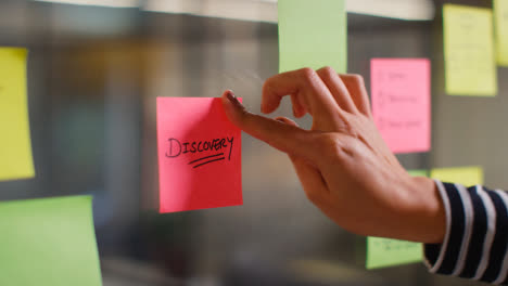 Close-Up-Of-Woman-Putting-Sticky-Note-With-Discovery-Written-On-It-Onto-Transparent-Screen-In-Office-2