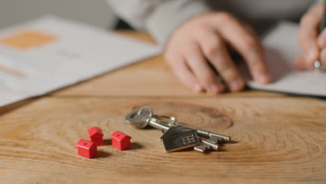 Hauskaufkonzept-Mit-Schlüsseln-Am-Schlüsselring-In-Hausform-Und-Person,-Die-Finanzen-überprüft-1