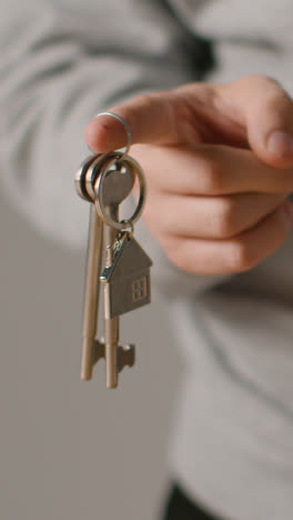 Vertical-Video-Home-Buying-Concept-With-Person-Holding-Keys-On-House-Shaped-Keyring-Against-Grey-Background