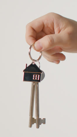 Vertical-Video-Home-Buying-Concept-With-Person-Holding-Keys-On-House-Shaped-Keyring-Against-White-Background