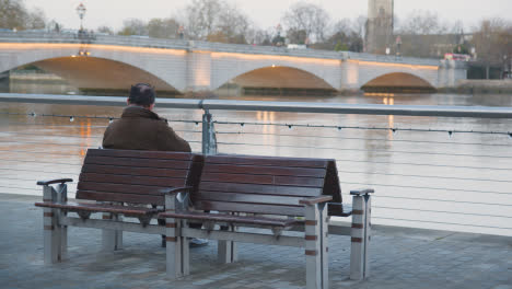 Man-Sitting-On-Bench-By-Putney-Bridge-Over-River-Thames-In-London-Illuminated-In-Winter