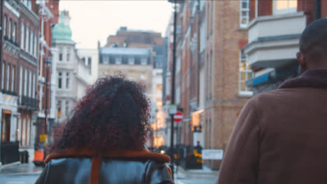 Rear-View-Of-Couple-Walking-Arm-In-Arm-Through-Street-On-Winter-Visit-To-London-