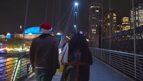 Vista-Trasera-De-Una-Pareja-Caminando-Sobre-El-Puente-Golden-Jubilee-En-Londres-Con-Sombreros-De-Navidad