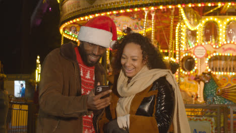 Couple-Celebrating-Christmas-Standing-By-Fairground-Roundabout-On-London-South-Bank-At-Night-With-Mobile-Phone