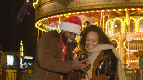 Couple-Celebrating-Christmas-Standing-By-Fairground-Roundabout-On-London-South-Bank-At-Night-With-Mobile-Phone-1