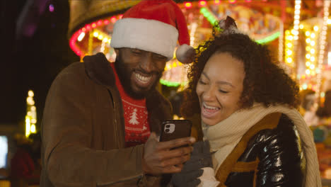 Couple-Celebrating-Christmas-Standing-By-Fairground-Roundabout-On-London-South-Bank-At-Night-With-Mobile-Phone-3