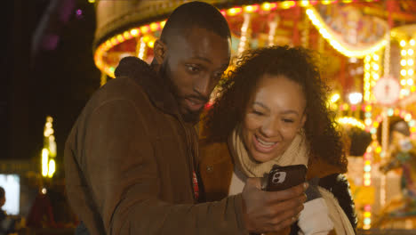 Couple-Standing-By-Fairground-Roundabout-On-London-South-Bank-At-Night-With-Mobile-Phone-3