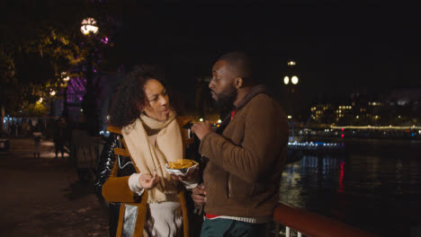 Couple-On-Date-Standing-On-London-South-Bank-At-Night-Eating-Chips