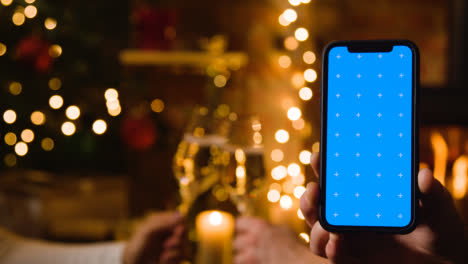 Paar-Macht-Weihnachtstoast-Mit-Champagner-Vor-Dem-Feuer-Zu-Hause-Mit-Handy-Mit-Blauem-Bildschirm