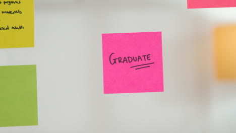 Close-Up-Of-Woman-Putting-Sticky-Note-With-Graduate-Written-On-It-Onto-Transparent-Screen-In-Office