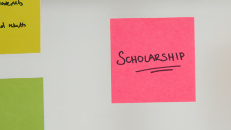 Close-Up-Of-Woman-Putting-Sticky-Note-With-Scholarship-Written-On-It-Onto-Transparent-Screen-In-Office-1