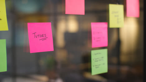 Close-Up-Of-Woman-Putting-Sticky-Note-With-Tutors-Written-On-It-Onto-Transparent-Screen-In-Office