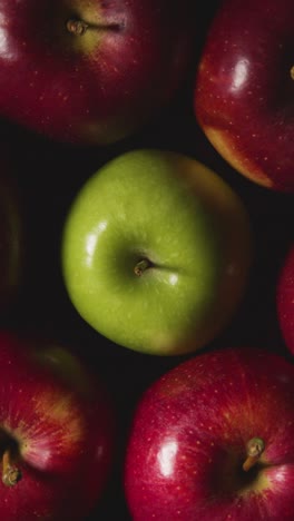 Vertical-Video-Overhead-Studio-Shot-Of-Green-Apple-Surrounded-by-Red-Apples-Revolving-Against-Black-Background