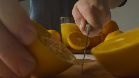 Cerca-Del-Hombre-Cortando-Naranjas-Con-Un-Vaso-De-Jugo-Fresco-En-Segundo-Plano.