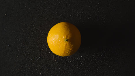 Obenliegende-Studioaufnahme-Von-Orange-Mit-Wassertröpfchen,-Die-Sich-Vor-Schwarzem-Hintergrund-Drehen
