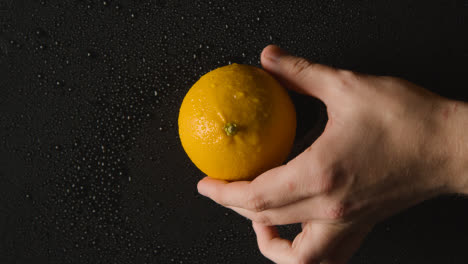 Overhead-Studioaufnahme-Einer-Hand,-Die-Orange-Mit-Wassertröpfchen-Aufhebt,-Die-Sich-Vor-Schwarzem-Hintergrund-Drehen