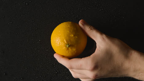 Foto-De-Estudio-Superior-De-La-Mano-Poniendo-Naranja-Con-Gotas-De-Agua-Girando-Sobre-Fondo-Negro