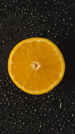 Vertical-Video-Overhead-Studio-Shot-Of-Orange-Half-With-Water-Droplets-Revolving-Against-Black-Background