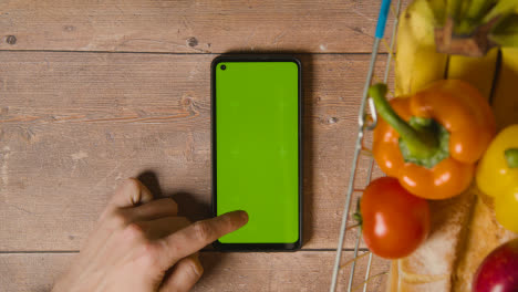Overhead-Studio-Shot-Of-Person-Using-Green-Screen-Mobile-Phone-Next-To-Basic-Food-Items-In-Supermarket-Wire-Shopping-Basket-