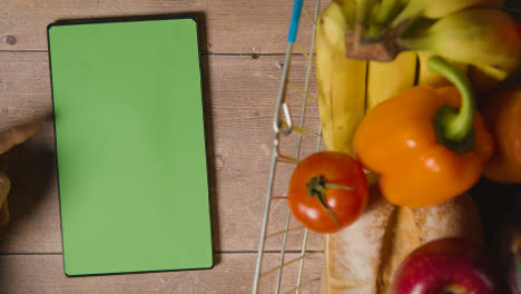 Overhead-Studio-Shot-Of-Person-Using-Green-Screen-Digital-Tablet-Next-To-Basic-Food-Items-In-Supermarket-Wire-Shopping-Basket-1