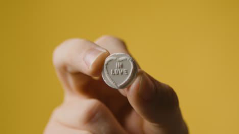 Close-Up-Of-Hand-Holding-Heart-Candy-With-In-Love-Message-On-Yellow-Background