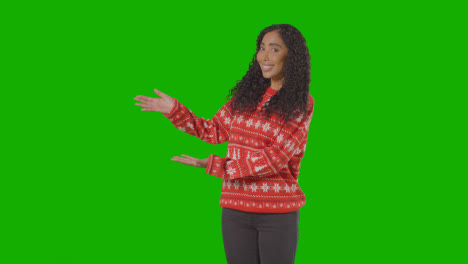Studioporträt-Einer-Frau,-Die-Einen-Weihnachtspullover-Trägt-Und-Einen-Gegenstand-Oder-Ein-Objekt-Vor-Einem-Grünen-Bildschirm-Zeigt,-Der-In-Die-Kamera-Lächelt