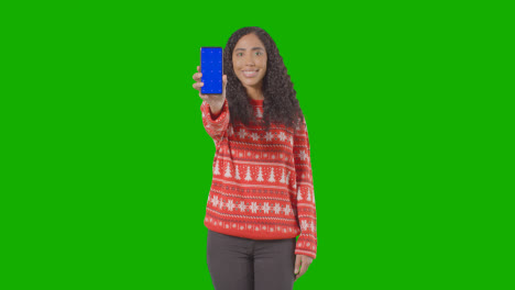 Studioporträt-Einer-Frau-Mit-Weihnachtspullover,-Die-Ihr-Handy-Gegen-Einen-Grünen-Bildschirm-Hochhält