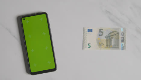 Overhead-Currency-Shot-Of-Hand-Grabbing-5-Euro-Note-Next-To-Green-Screen-Mobile-Phone