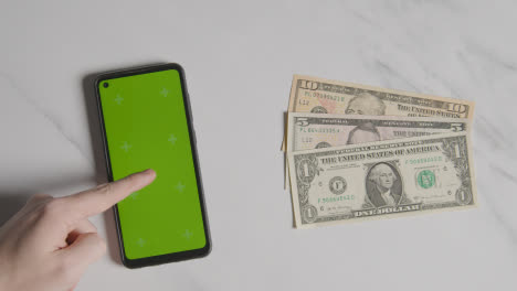 Overhead-Currency-Shot-Of-US-Dollar-Bills-Next-To-Person-Using-Green-Screen-Mobile-Phone