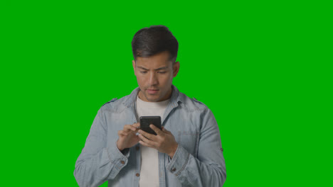 Studio-Shot-Of-Casually-Dressed-Young-Man-Online-Swiping-On-Mobile-Phone-Against-Green-Screen-