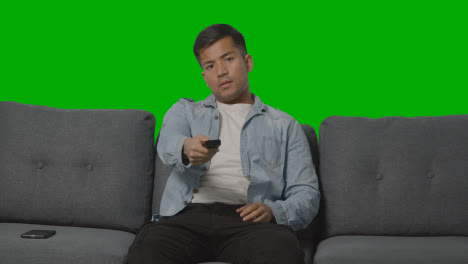 Studio-Shot-Of-Man-Looking-At-Mobile-Phone-Before-Sitting-On-Sofa-With-Remote-Control-Flicking-Through-TV-Channels-Against-Green-Screen-