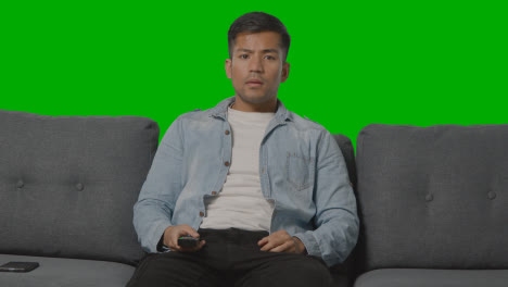 Studio-Shot-Of-Young-Man-Sitting-On-Sofa-With-Remote-Control-Flicking-Through-TV-Channels-Against-Green-Screen-