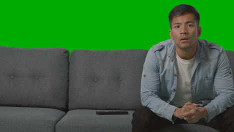 Studio-Shot-Of-Disappointed-Young-Man-Sitting-On-Sofa-Watching-Sport-On-TV-Against-Green-Screen-