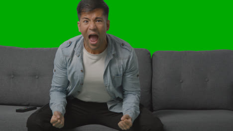 Studio-Shot-Of-Young-Man-Celebrating-Sitting-On-Sofa-Watching-Sport-On-TV-Against-Green-Screen-