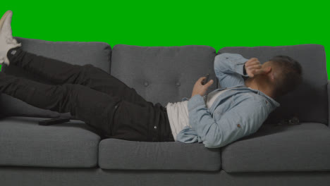 Young-Man-Lying-On-Sofa-Sneezing-And-Looking-At-Mobile-Phone-Against-Green-Screen